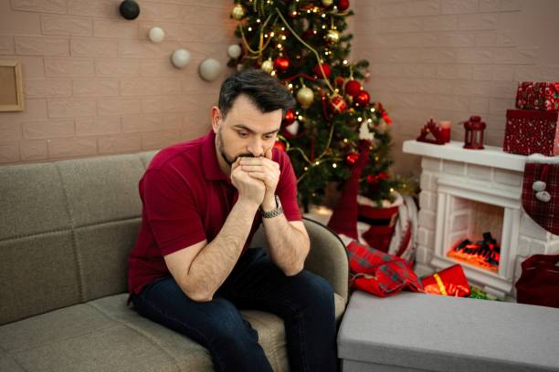istock Sad Young Man Sitting on Sofa Near Christmas Tree and Fireplace: Feeling Lonely During Holiday Season 2199448013
