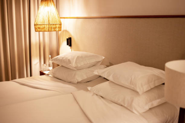 A stylish bedroom interior featuring a contemporary glass pendant light hanging beside a bed with crisp white pillows and a wooden headboard. The soft ambient lighting creates a cozy and elegant atmosphere. A beige curtain and a plush armchair complement the warm wooden tones, emphasizing a modern and minimalist design. Ideal for themes related to interior decor, hotel design, luxury bedrooms, Scandinavian aesthetics, and warm lighting concepts.
