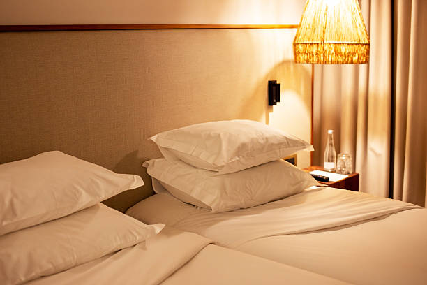A stylish bedroom interior featuring a contemporary glass pendant light hanging beside a bed with crisp white pillows and a wooden headboard. The soft ambient lighting creates a cozy and elegant atmosphere. A beige curtain and a plush armchair complement the warm wooden tones, emphasizing a modern and minimalist design. Ideal for themes related to interior decor, hotel design, luxury bedrooms, Scandinavian aesthetics, and warm lighting concepts.