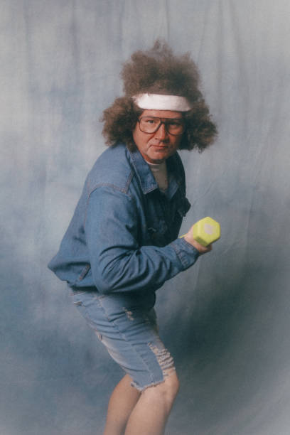 A man with a denim jacket sports a large curly hairstyle with sweatband and oversized glasses, posing for some ironic glam shots in vintage kitsch style.  He does bicep curls with a small dumbbell weight.  Soft focus in style of mall professional portrait styles.