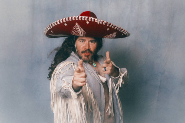 A man with a fringe lined jean jacket and oversized sombrero hat poses for some ironic glam shots in vintage kitsch style.  He points his fingers in a pretend shooting a gun gesture.  Soft focus in style of mall professional portrait styles.