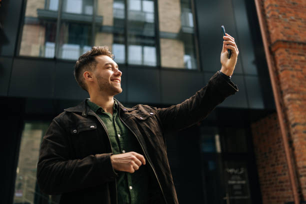 istock Low-angle view of cheerful handsome man holding smartphone in hand making video call, standing in front of modern buildings in city center. 2199471007