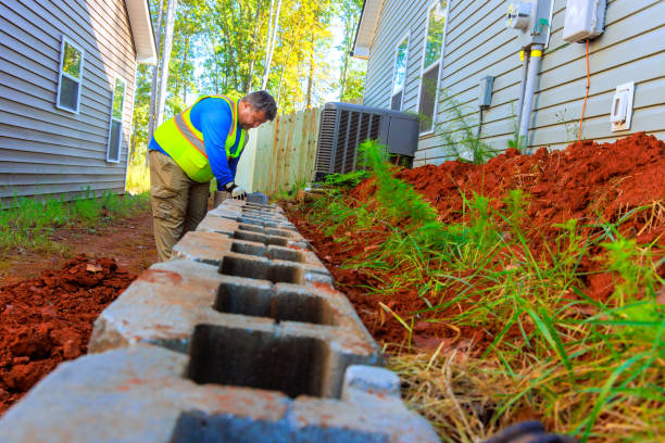 Worker lays foundation blocks beside residential building during sunny afternoon in a suburban area
