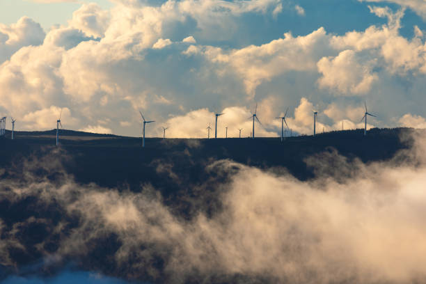 Wind turbines stand tall on a misty mountain ridge, partially veiled by low-hanging clouds. Dramatic sky, filled with billowing clouds, enhances contrast between nature and renewable energy technology