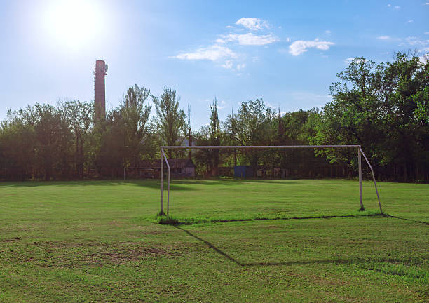 Sunlit soccer field with a rusty goalpost stands surrounded by lush green trees, evoking nostalgia for casual games. In background an old industrial chimney rises above the treetops