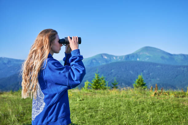 istock Woman stands in grassy meadow, using binoculars to observe distant mountains. 2199486372