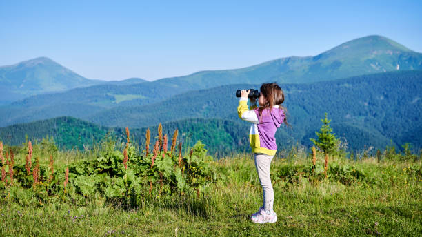 Young girl standing outdoors looking through binoculars, exploring trees and fallen leaves in a vibrant autumn park