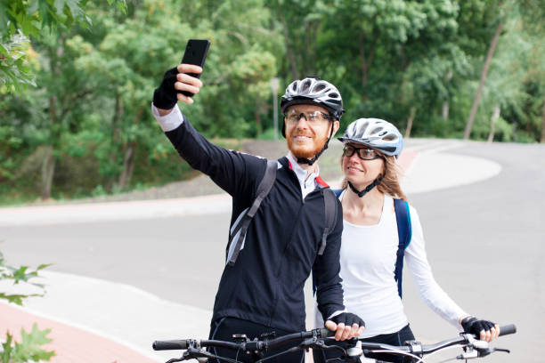 istock Cyclists take selfies, using mobile phone on bicycle path. Happy couple in protective helmets having fun on road. Healthy lifestyle. Sports activity outdoors in nature. Local ecological travel 2199490548