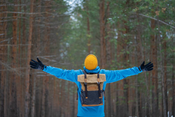 Hiker with backpack and yellow hat enjoying freedom with open arms in beautiful winter forest, embracing the tranquility of nature during snowfall