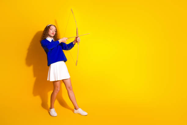 istock Young woman posing with a bow and arrow on a vibrant yellow background 2199554504