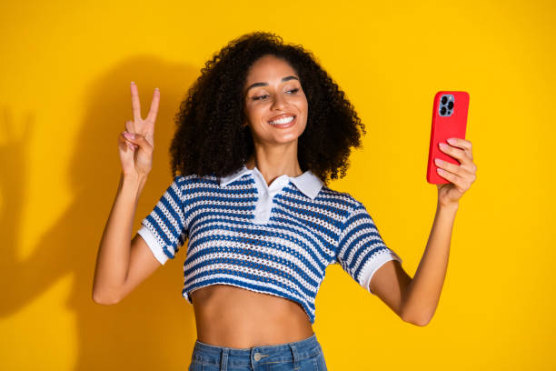 istock Stylish young woman taking a cheerful selfie on a yellow background while making a peace sign, wearing a striped top 2199558836