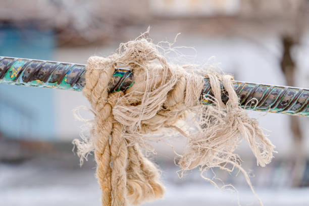 A close-up of an old, frayed rope tightly knotted around a weathered metal bar. The fibers of the rope are unraveling, giving it a rugged, textured appearance. The metal bar shows signs of wear with chipped paint and rust, highlighting its age and exposure to the elements. The background is blurred, drawing attention to the intricate details of the rope and bar.