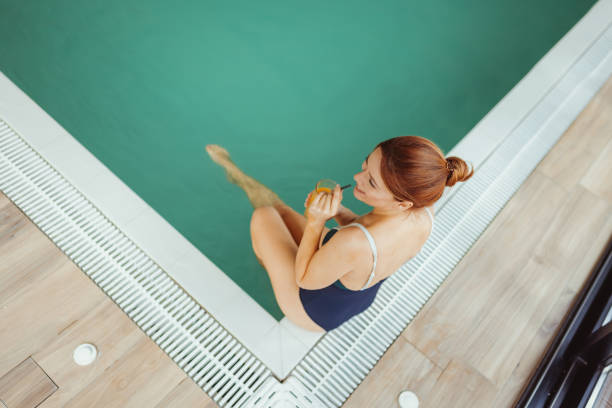 A Content Woman Sits at the Edge of a Serene Spa Pool, Sipping on a Refreshing Juice. Her Relaxed Posture and Bright Smile Embody the Perfect Spa Experience, Ideal for Wellness, Relaxation, and Self-care Marketing.