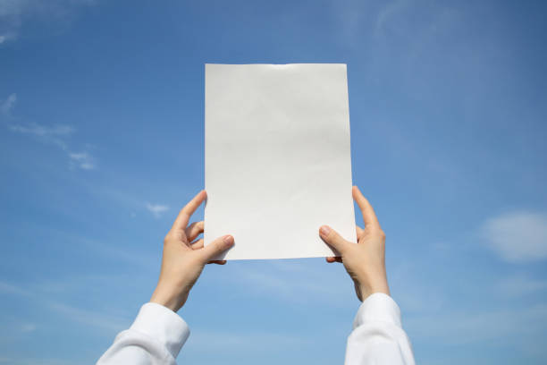 istock Hands holding up a blank sheet of paper outdoors, on a blue sky background. 2199587314