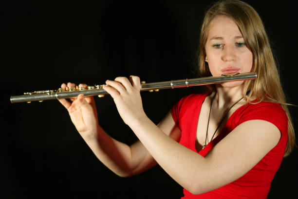 A young blonde woman with long hair, wearing a red T-shirt, practicing playing the flute against a black background. Front lighting