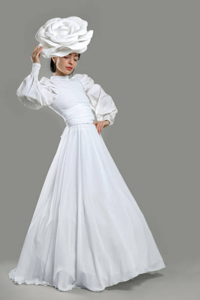 A woman in a long white dress and a hat in the form of a white flower poses on a gray background, creating an elegant and sophisticated image.