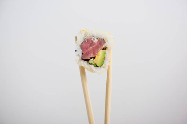 Fresh tuna sushi California roll with avocado and sesame seeds is delicately held by chopsticks against a clean white background. Minimalist and appetizing.