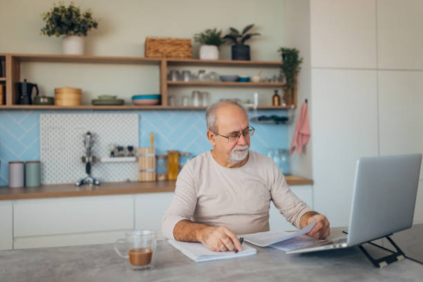 istock Concentrated senior man with eyeglasses uses his laptop for further education through e-learning. He sits comfortably at the kitchen table, taking advantage of technology to advance his knowledge 2199623426