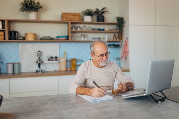 istock Smiling senior man sitting at kitchen counter and using laptop for online education and training course 2199623430