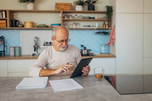 istock Senior man with eyeglasses uses his digital tablet for further education through e-learning. He sits comfortably at the kitchen table, taking advantage of technology to advance his knowledge 2199623641