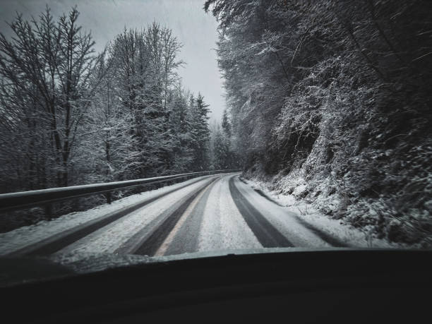 A first-person perspective of driving on a winding road covered in fresh snow, surrounded by a dense winter forest. The icy conditions and tire tracks on the road highlight the challenges of winter travel, creating a moody and atmospheric scene. Ideal for themes related to road trips, seasonal changes, and winter safety.