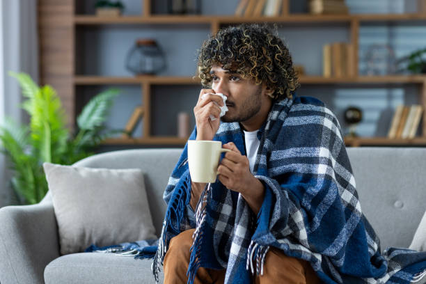 istock Sick man with a cold sits at home on the couch, covered with a blanket, and with a napkin in his hand. A man with a cold with a hot drink in his hands has a runny nose 2199632793