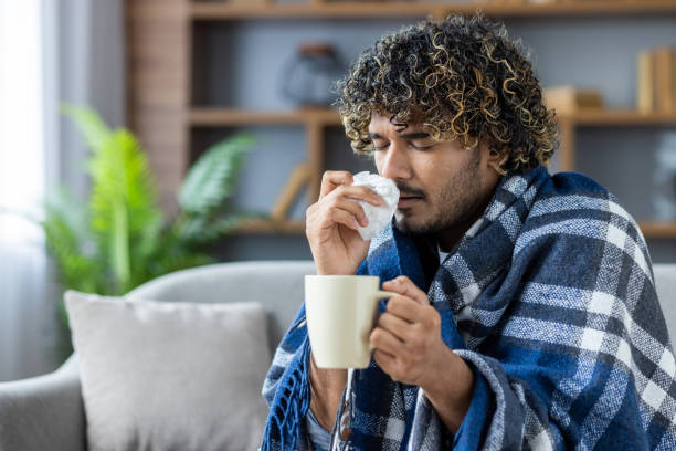istock Man battling cold wrapped in blanket holding cup and tissue indoors 2199632794