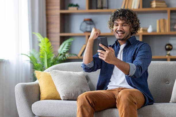 Successful joyful man sitting at home on sofa with phone in hands, celebrating victory and successful achievement results. Freelancer received online notification with good news on smartphone