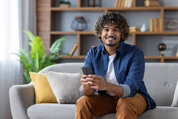istock Young man with phone in hands smiling and looking at camera, using app on smartphone, sitting on sofa in living room of apartment at home 2199633508