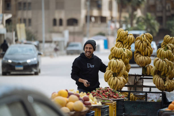 Giza, Egypt - January 23, 2025: Day at the local groceries market on the streets of Giza, famous for historic landmarks - Giza Pyramids. Cheerful fruit salesman attracting customers with the smile.