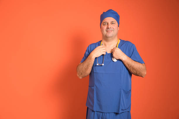 Confident mature doctor in blue scrubs and surgical cap adjusting a yellow stethoscope while smiling at the camera against a vibrant orange background