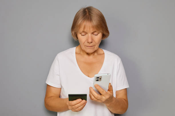 istock A middle-aged woman stands in a plain room while examining her smartphone and holding a bank card in her other hand, possibly considering a purchase online or managing finances 2199644730