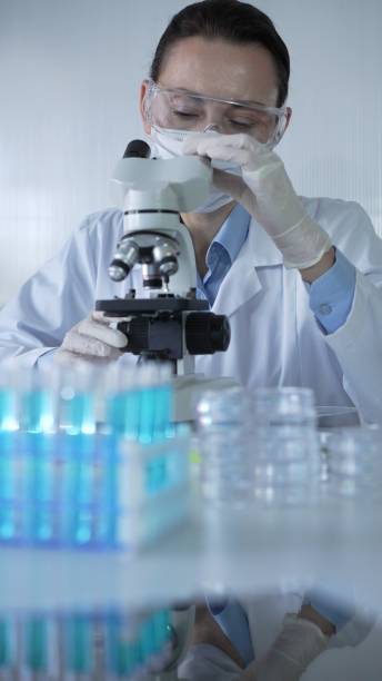 istock Woman scientist wearing lab coat, white protective gloves and glasses, is using microscope in modern laboratory setup, portrait view. Low key lighting photo. Science and Medicine concept 2199677877