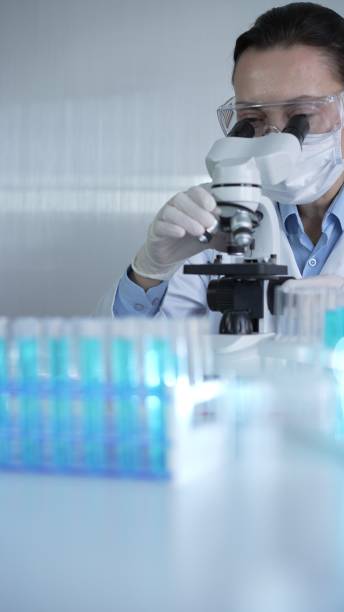 istock Woman scientist wearing lab coat, white protective gloves and glasses, is using microscope in modern laboratory setup, portrait view. Low key lighting photo. Science and Medicine concept 2199681131