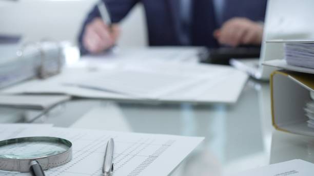 istock Magnifying glass, calculator, pen, laptop and documents are lying on accountant's desk. Businessman wearing blue suit is working with documents on the background. Business people and audit concept 2199681232