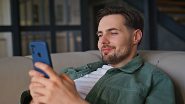 istock Closeup guy messaging smartphone lying at light couch. Relaxed happy man online 2199686649