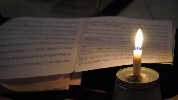 A burning candle on a metal surface illuminates an open sheet of music in a dimly lit environment, creating a nostalgic and intimate atmosphere.