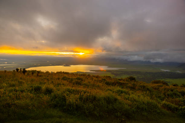 A serene sunset blankets a Tanzanian landscape with a lake reflecting the warm glow, surrounded by lush greenery under a dramatic sky.