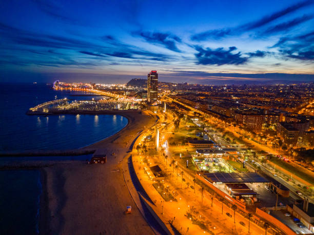 A sweeping twilight shot of Barcelona displays the lit coastline, prominent buildings, and serene beach, as the city transitions from dusk to night.