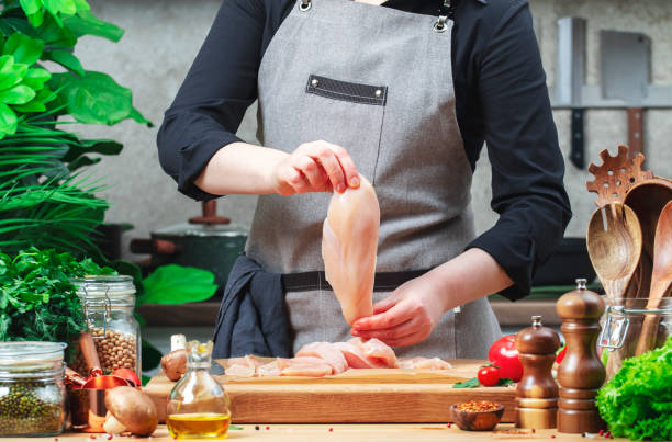 istock Chef holding raw chicken breast fillet in hands. Cozy kitchen with wooden table, kitchenware, vegetables, herbs and ingredients for cooking. Healthy eating, culinary, recipes, food blogging 2199754465