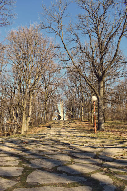 A scenic stone pathway leads through a forest toward the Kosmaj Monument, a WWII memorial in Serbia. The brutalist structure stands tall in the distance, framed by leafless trees and lampposts.