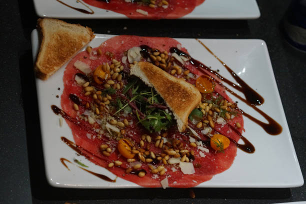 A delicious plate of beef carpaccio is artfully arranged with pine nuts, shavings of cheese, fresh greens, and a drizzle of balsamic glaze, served with toasted bread.
