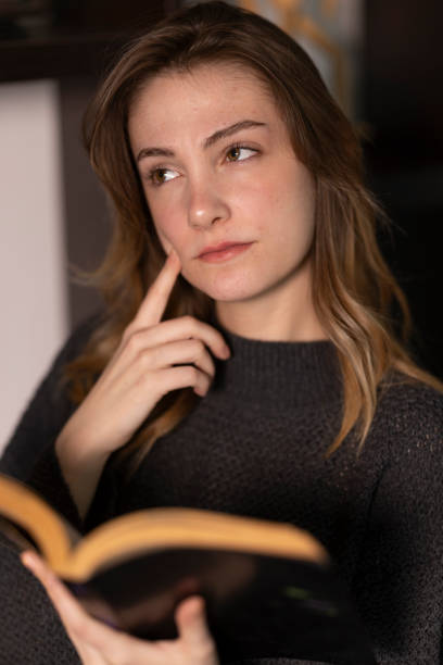 Thoughtful young woman reading a book at home, touching her face with her finger