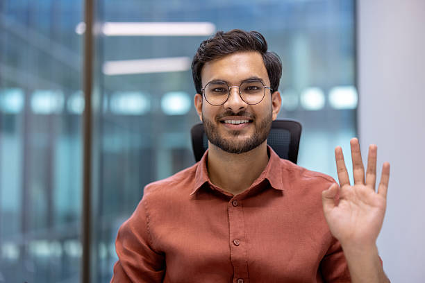 istock Confident young businessman waving and smiling in modern office setting, demonstrating approachable and friendly demeanor ideal for corporate and professional environments 2199803544