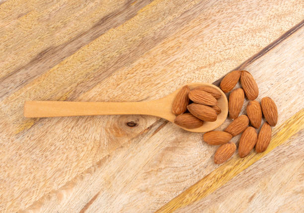Almonds in spoon pile, nut group, whole almond seeds, brown scattered fruit kernels, dry raw nuts on wood background top view