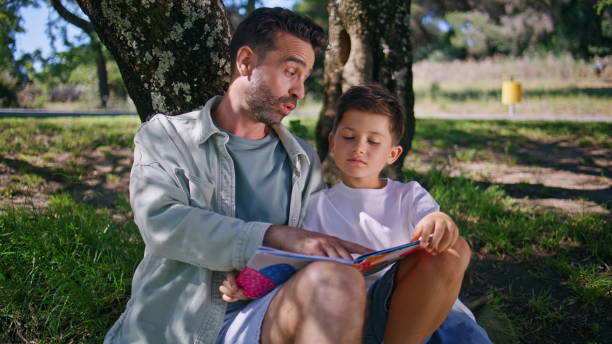 istock Cute family reading book sitting grass at forest closeup. Father son under tree 2199819074