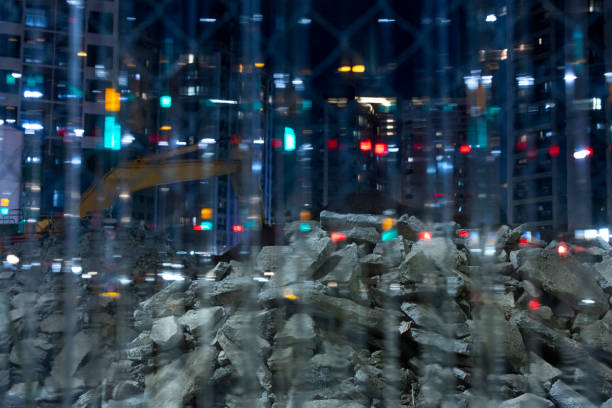 A construction site with rocks in the city at night, mixed with blurred lights and trailing shadows, with an excavator under construction and city buildings in the background.