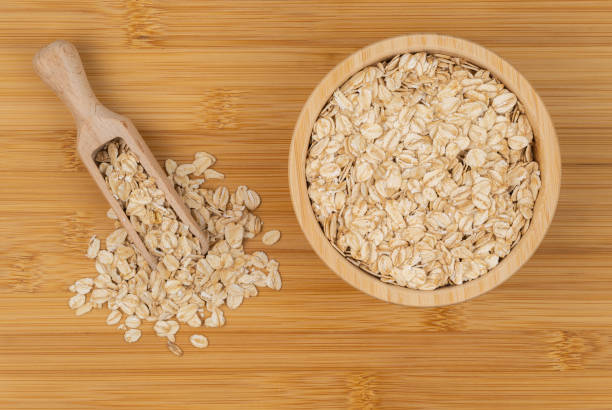 A wooden bowl filled with rolled oats alongside a wooden scoop spilling oats onto a bamboo surface. The natural texture and warm tones highlight the wholesome and organic quality of the grains.