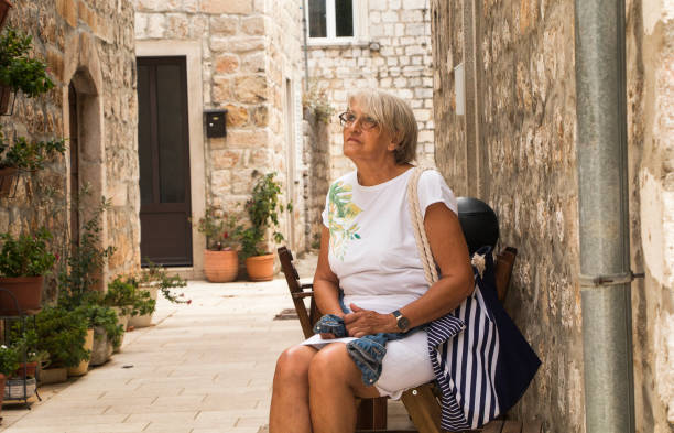 A woman with gray hair is seated on a bench, dressed in light clothing, looking relaxed and thoughtful. The backdrop features a contemporary geometric wall made of metal panels.