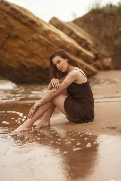 brunette girl is sitting on a sandy beach. the girl has long hair she is wearing a thin black dress. around her are big yellow rocks and the sea. her dress is wet, the girl is thoughtful.